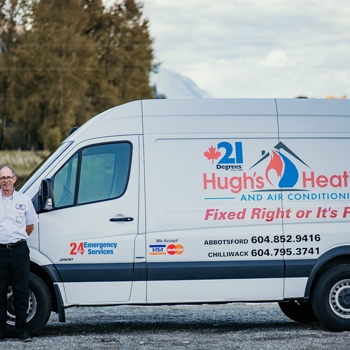 A person stands next to a white 21 Degrees Hugh's Heating and Air Conditioning truck with tall trees in the background